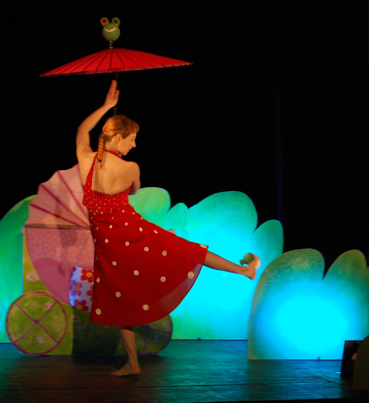 Spectacle Rosie Rose Baby Karen Bourre en robe rouge à pois blancs jongle avec une ombrelle rouge et deux grenouilles vertes devant le décors de parc vert